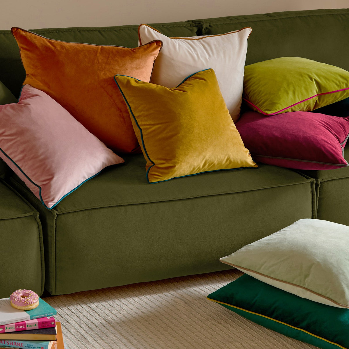 Meridian Plain Velvet Cushion in ecru with ginger piped edge, shown among an assortment of coloured velvet scatter cushions on a green sofa.