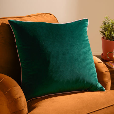 Meridian velvet cushion in emerald green with a blush pink piped edge, shown square on an orange velvet sofa beside a potted plant.
