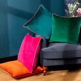 Meridian velvet cushion in emerald green with blush pink piped edge, shown on a dark grey armchair alongside teal, pink, and orange velvet cushions.