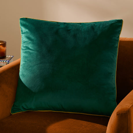 Meridian Plain Velvet Cushion in emerald green with contrasting piped edge, square velvet-feel design shown on a rust-coloured armchair.
