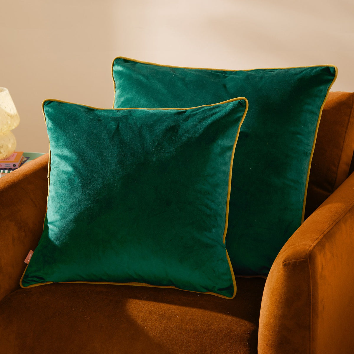 Two Meridian plain velvet cushions in emerald green with contrasting moss piped edges, styled on a burnt orange velvet armchair.