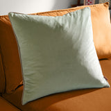 Meridian Plain Velvet Cushion in sage green, a square velvet cushion with taupe piped trim edging, shown propped on a mustard-orange sofa.