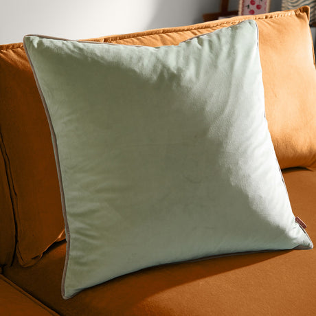 Meridian Plain Velvet Cushion in sage green, a square velvet cushion with taupe piped trim edging, shown propped on a mustard-orange sofa.