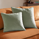 Two Meridian sage green velvet cushions with taupe piped trim, styled on an orange sofa in a living room setting.
