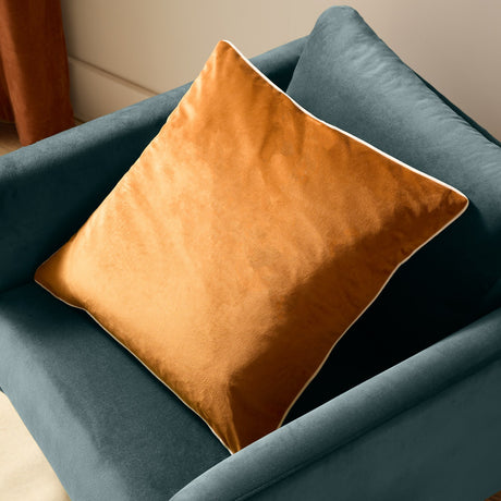 Meridian Plain Velvet Cushion in ginger orange with contrasting ecru piped edge, shown square on a teal velvet armchair.