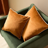 Two Meridian square velvet cushions in ginger with contrasting ecru piped edges, styled on a green velvet armchair.