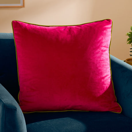 Meridian Plain Velvet Cushion in hot pink with lime green piped edge, shown on a blue velvet sofa in a living room setting.