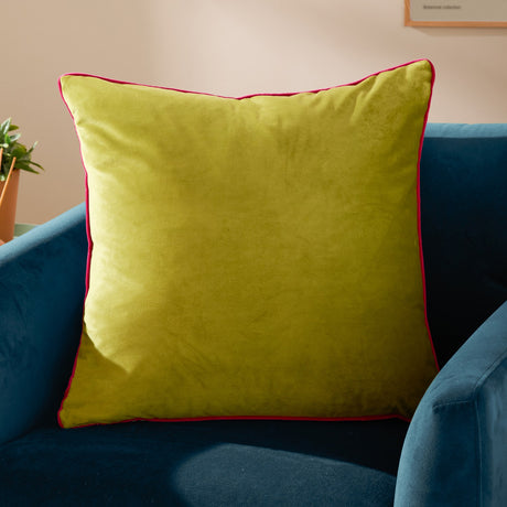 Meridian Plain Velvet Cushion in lime green with hot pink piped edging, shown on a blue velvet armchair in a living room setting.