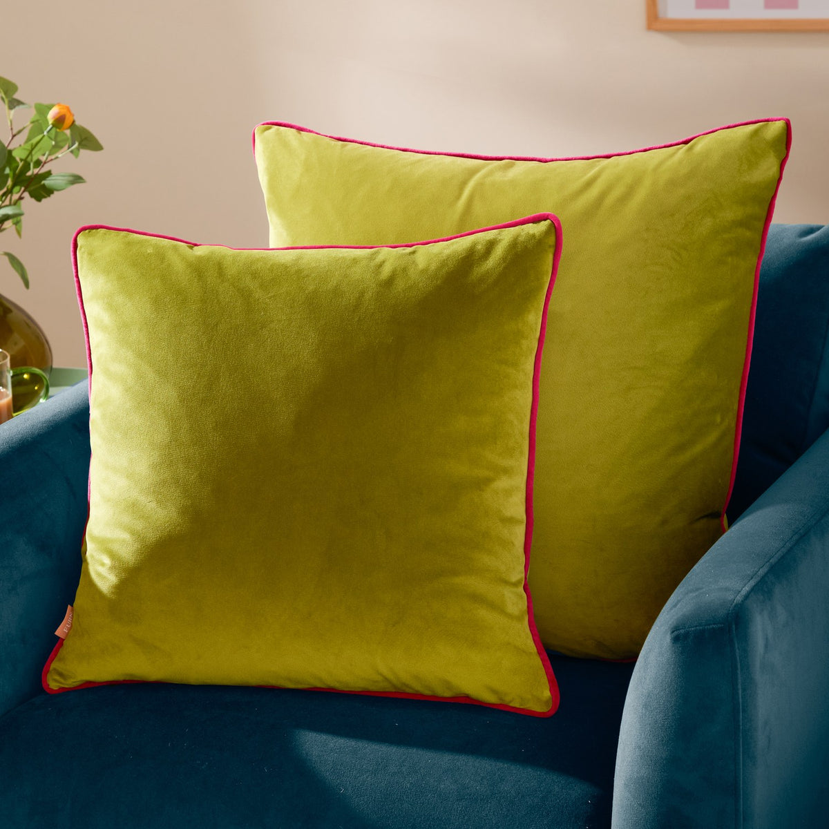 Two Meridian square velvet cushions in lime green with contrasting hot pink piped edges, styled on a teal velvet armchair.