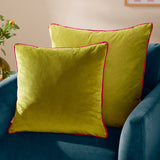 Two Meridian square velvet cushions in lime green with contrasting hot pink piped edges, styled on a teal velvet armchair.