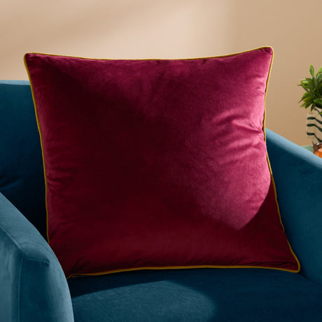 Meridian Plain Velvet Cushion in maroon with moss green piped edging, shown on a teal armchair against a neutral wall.