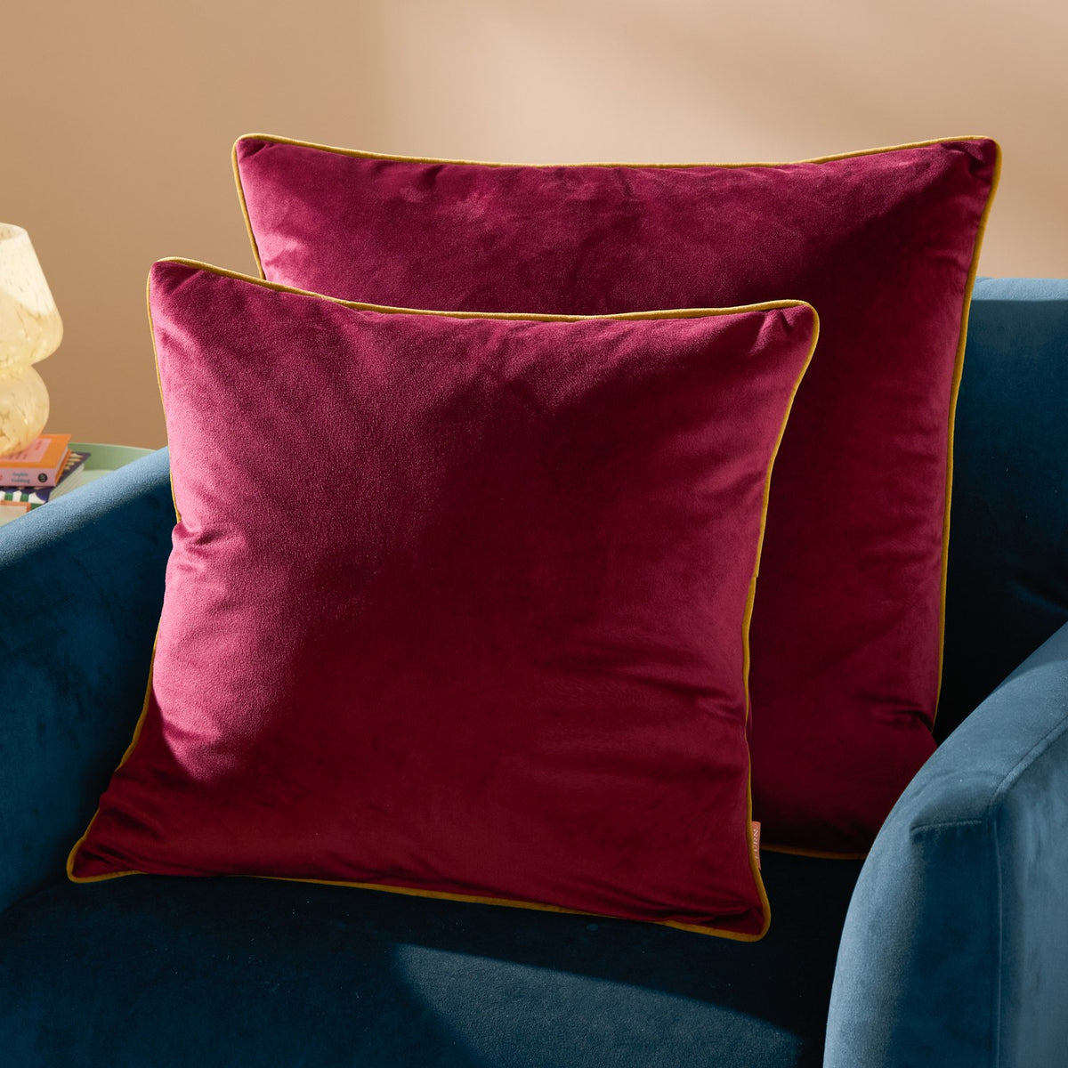 Meridian Plain Velvet Cushions in maroon with moss green piped edging, shown styled on a teal armchair against a warm neutral background.
