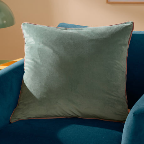 Meridian Plain Velvet Cushion in mineral blue with blush pink piped edge, square 55x55cm, shown on a teal velvet sofa.