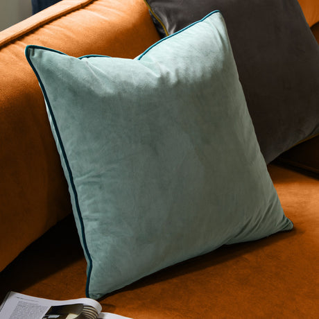 Meridian Plain Velvet Cushion in mineral blue with contrasting teal piped edge, shown on a burnt orange sofa alongside a dark grey velvet cushion.