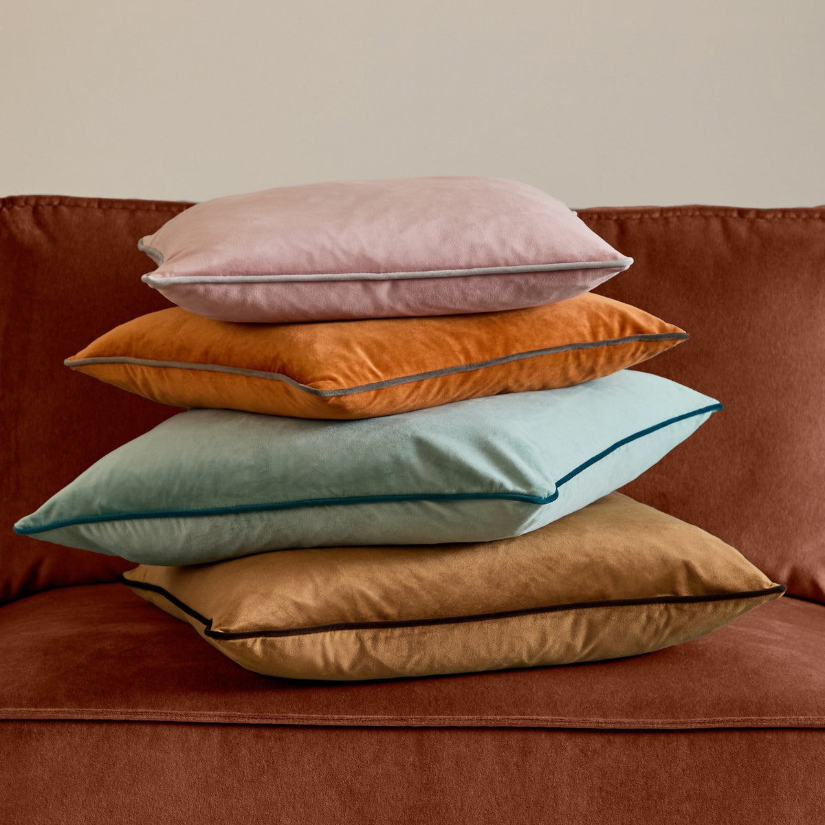 Meridian Plain Velvet Cushion in mineral blue with teal piped edge, shown third from top in a stacked set of four velvet cushions on a rust sofa.