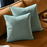 Two Meridian square velvet cushions in mineral blue with contrasting teal piped edges, styled on a tan sofa beside a wooden tray.