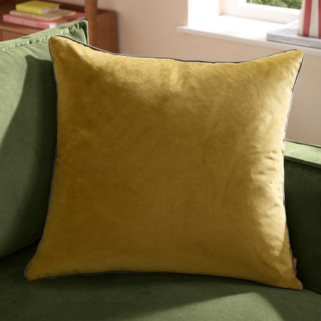 Meridian Plain Velvet Cushion in mustard yellow with charcoal piped edging, shown on a green sofa in a living room setting.