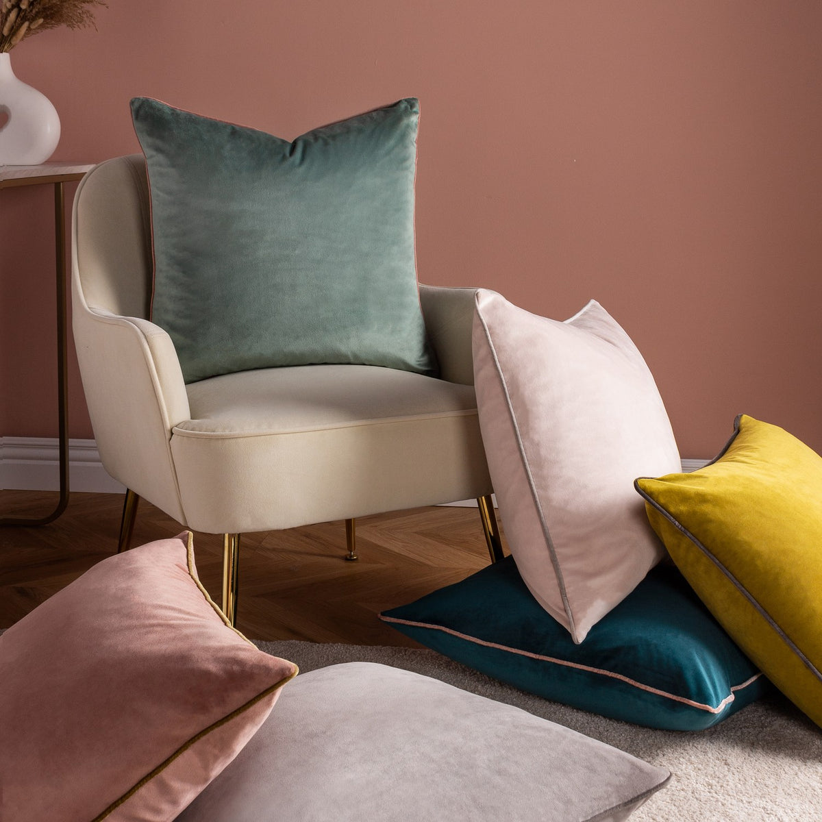 Meridian Plain Velvet Cushion in moss green with charcoal piped edges, shown on a cream armchair alongside pink, grey, teal and yellow velvet cushions in a living room setting.