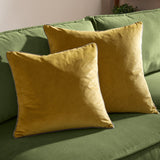 Two square Meridian velvet cushions in mustard-yellow with grey piped edges, styled on a green sofa.