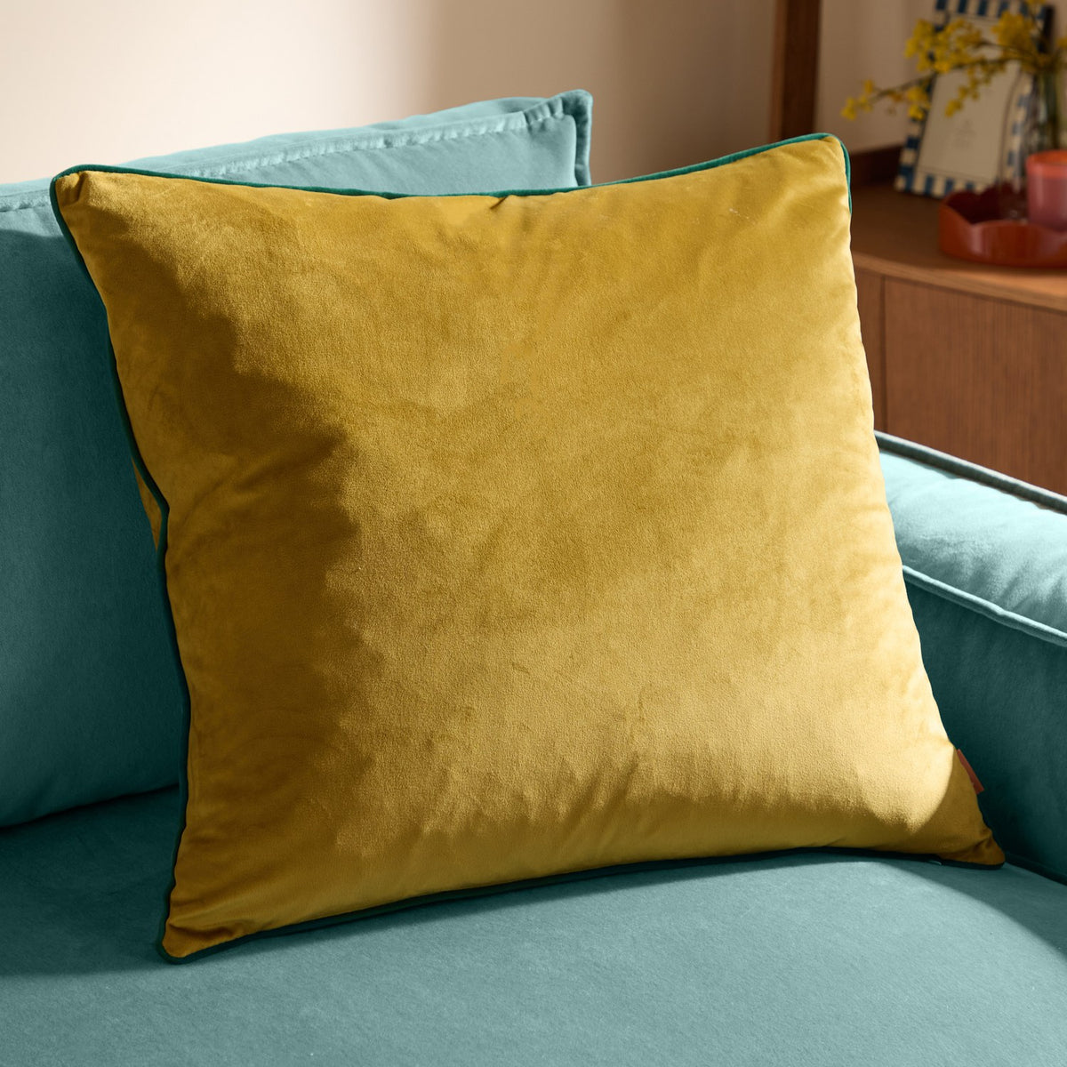 Meridian Plain Velvet Cushion in mustard-gold velvet with emerald piped edging, shown square-shaped on a teal sofa in a living room setting.