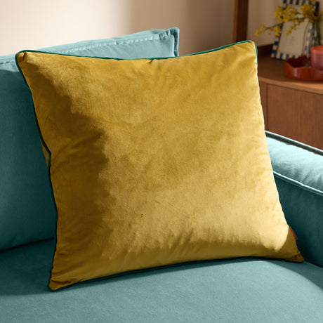 Meridian Plain Velvet Cushion in mustard-gold velvet with emerald piped edging, shown square-shaped on a teal sofa in a living room setting.