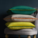 Stack of three velvet cushions on a grey armchair, with the top emerald green cushion featuring a contrasting yellow piped border, above a taupe and a yellow cushion.