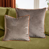 Two square Meridian velvet cushions in mocha brown with pumpkin orange piped edges, styled on a green velvet sofa near a curtained window.