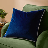 Meridian Plain Velvet Cushion in navy blue with silver piped edging, shown on a green sofa arm beside a plant and mug on a side table.