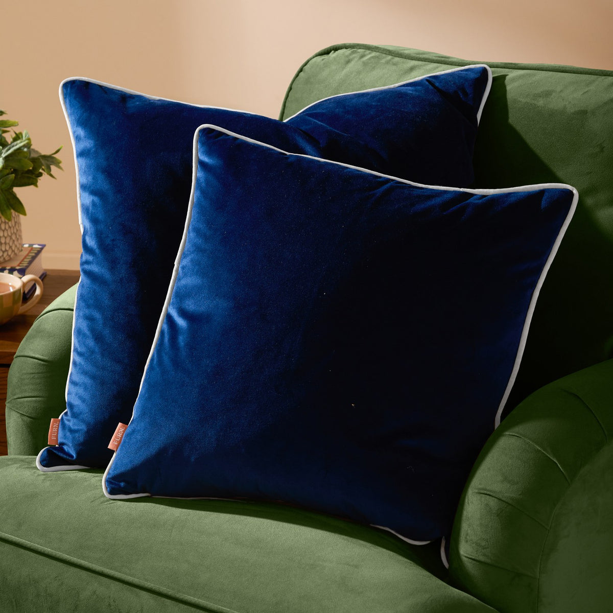 Two Meridian Plain Velvet Cushions in navy blue with contrasting silver piping, styled on a green velvet sofa.