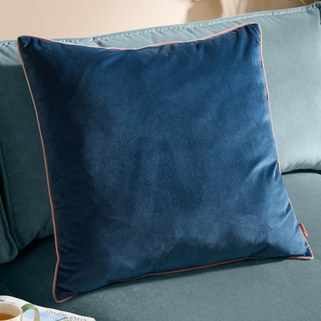 Meridian Plain Velvet Cushion in petrol blue with blush pink piped edging, shown on a teal sofa in a living room setting.