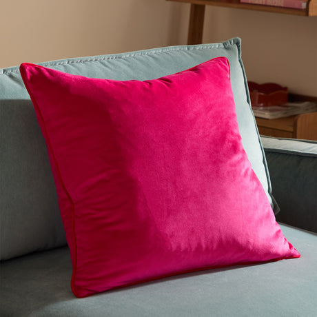 Meridian plain velvet cushion in hot pink/magenta with contrasting red piped edging, shown on a light grey sofa in a living room setting.