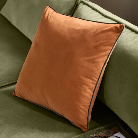 Meridian Plain Velvet Cushion in pumpkin orange with mocha brown piped edging, shown propped on a green sofa in a living room setting.