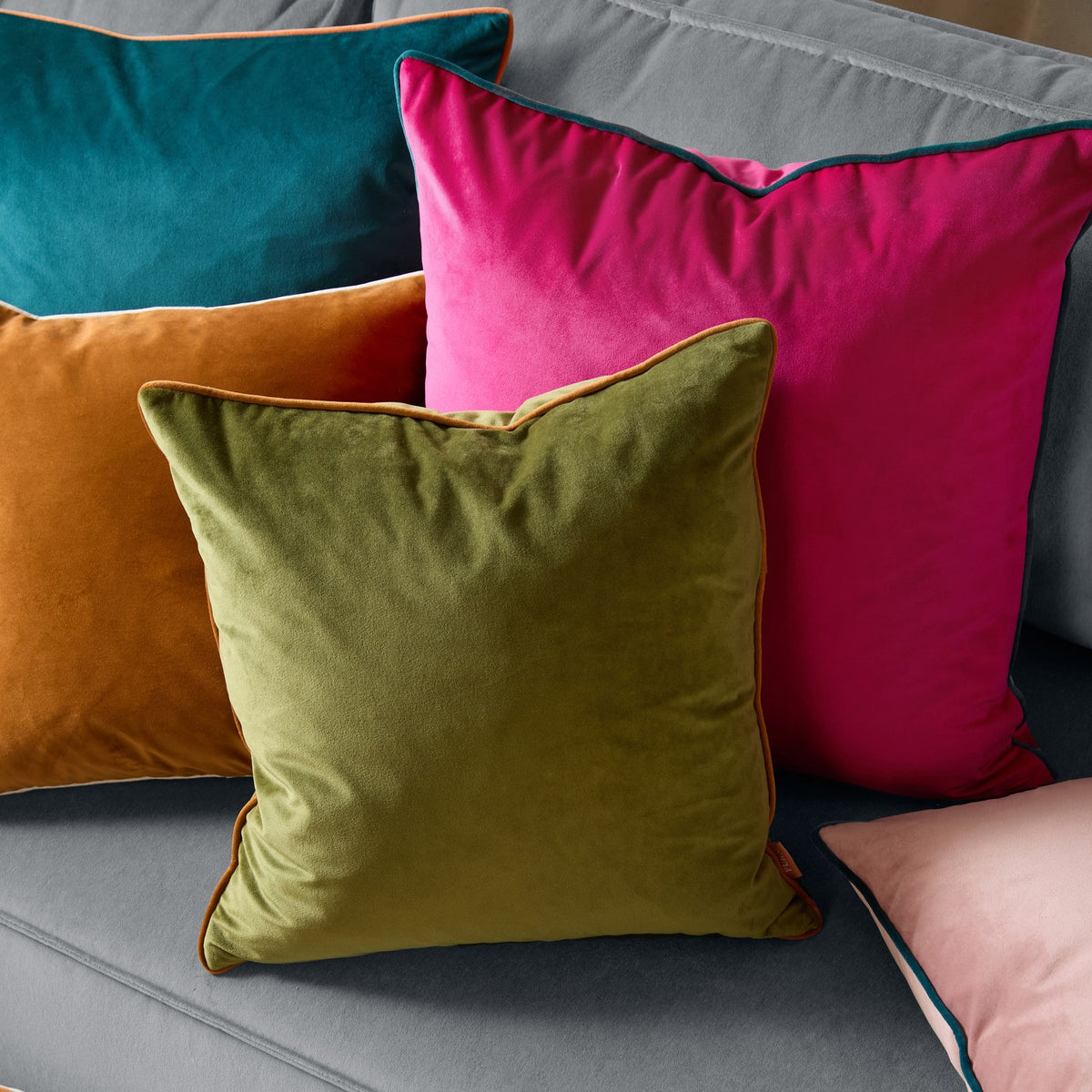 Meridian Plain Velvet Cushion in raspberry pink with contrasting teal piped edge, shown among olive, orange and teal velvet cushions on a grey sofa.