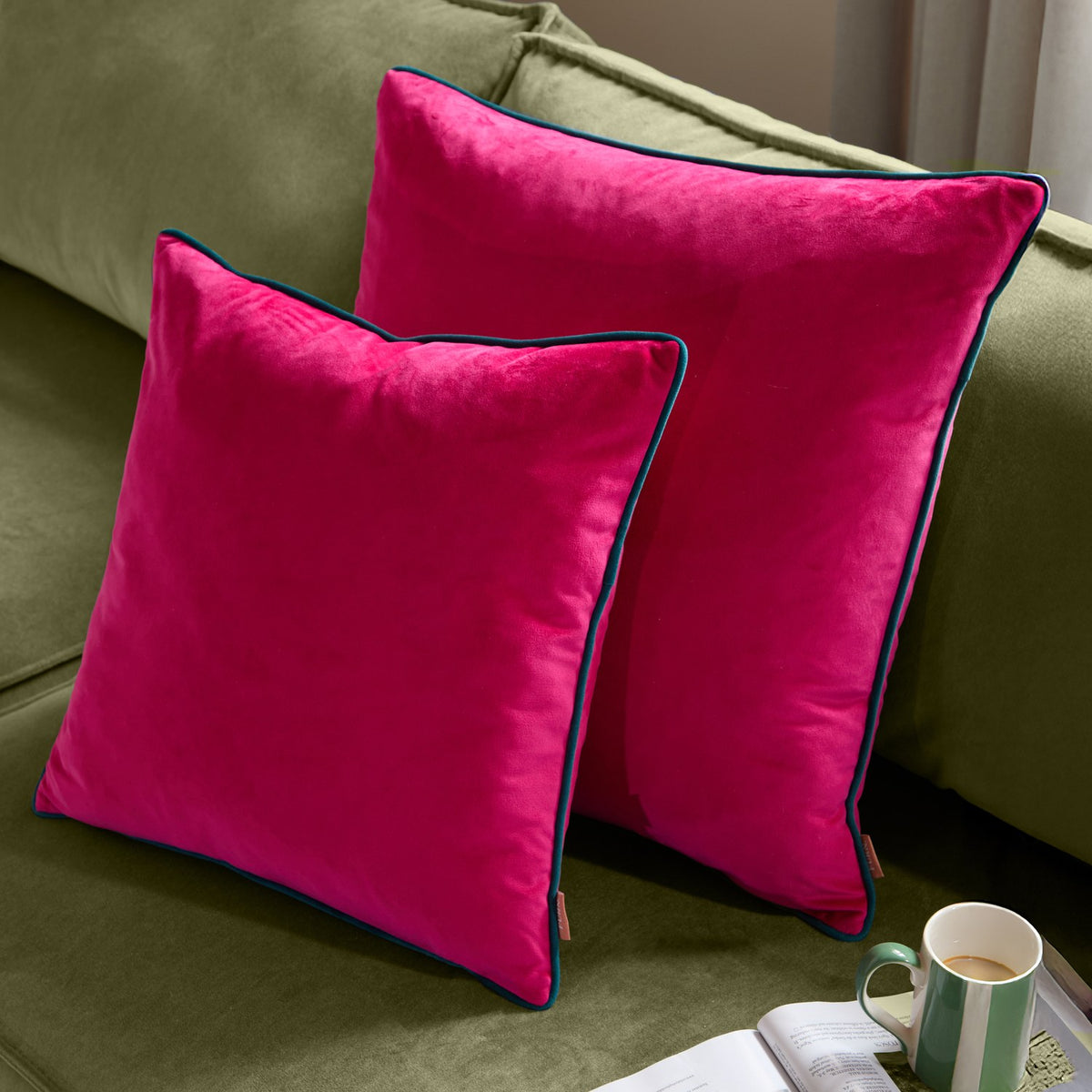 Two Meridian square velvet cushions in raspberry pink with contrasting teal piped edges, shown on an olive green sofa in a living room setting.