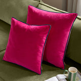 Two Meridian square velvet cushions in raspberry pink with contrasting teal piped edges, shown on an olive green sofa in a living room setting.