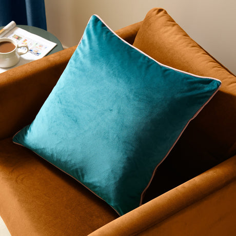 Meridian Plain Velvet Cushion in deep teal with blush pink piped edging, shown on a rust velvet armchair in a living room setting.