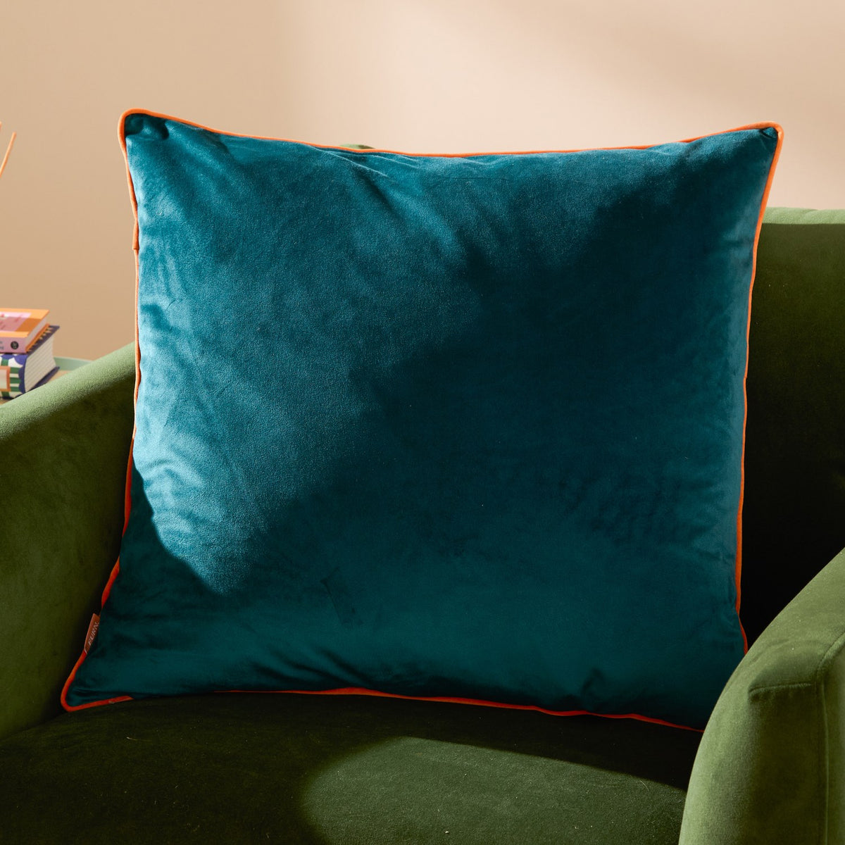 Meridian Plain Velvet Cushion in teal with clementine orange piped edging, shown square-shaped on a green upholstered armchair.