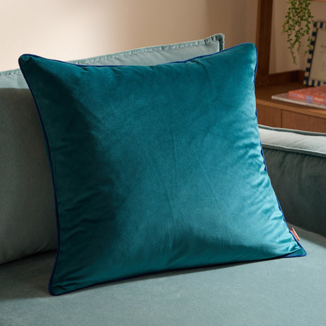 Meridian Plain Velvet Cushion in deep teal with contrasting navy piped edging, shown resting on a light green sofa in a living room setting.