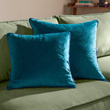 Two Meridian Plain Velvet Cushions in teal with contrasting navy piped edges, styled on a green sofa in a living room setting.