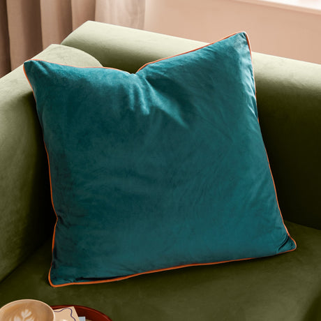Meridian Plain Velvet Cushion in deep teal with contrasting orange piped edge, shown propped on a green sofa in a living room setting.