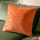 Meridian Plain Velvet Cushion in tiger orange with contrasting teal piped edge, shown on a green sofa in a living room setting.