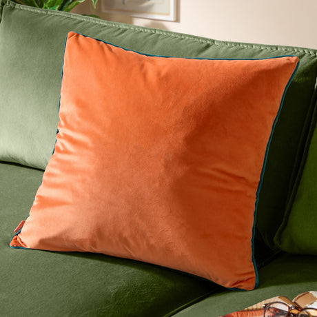 Meridian Plain Velvet Cushion in tiger orange with contrasting teal piped edge, shown on a green sofa in a living room setting.