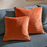 Two square Meridian velvet cushions in tiger orange with contrasting teal piped edges, shown on a blue upholstered sofa.