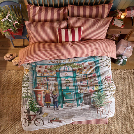 Merry & Bright cotton striped duvet cover set on a bed, showing the reversible bakery shopfront print with striped pink and red pillowcases in a Christmas-decorated bedroom.