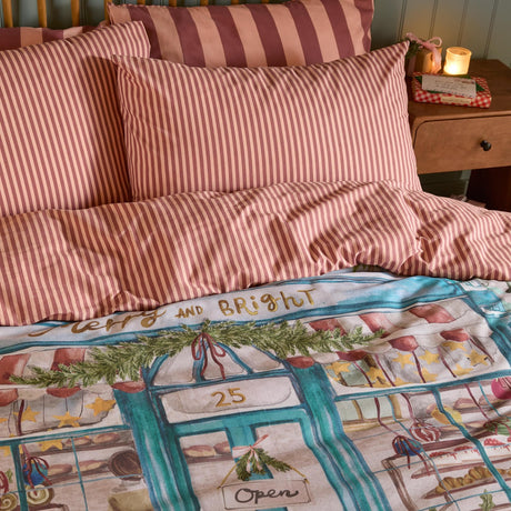 Merry & Bright Cotton Striped Duvet Cover Set in pink and red candy stripe, styled on a bed with matching striped pillowcases and a glimpse of the reverse bakery scene print.