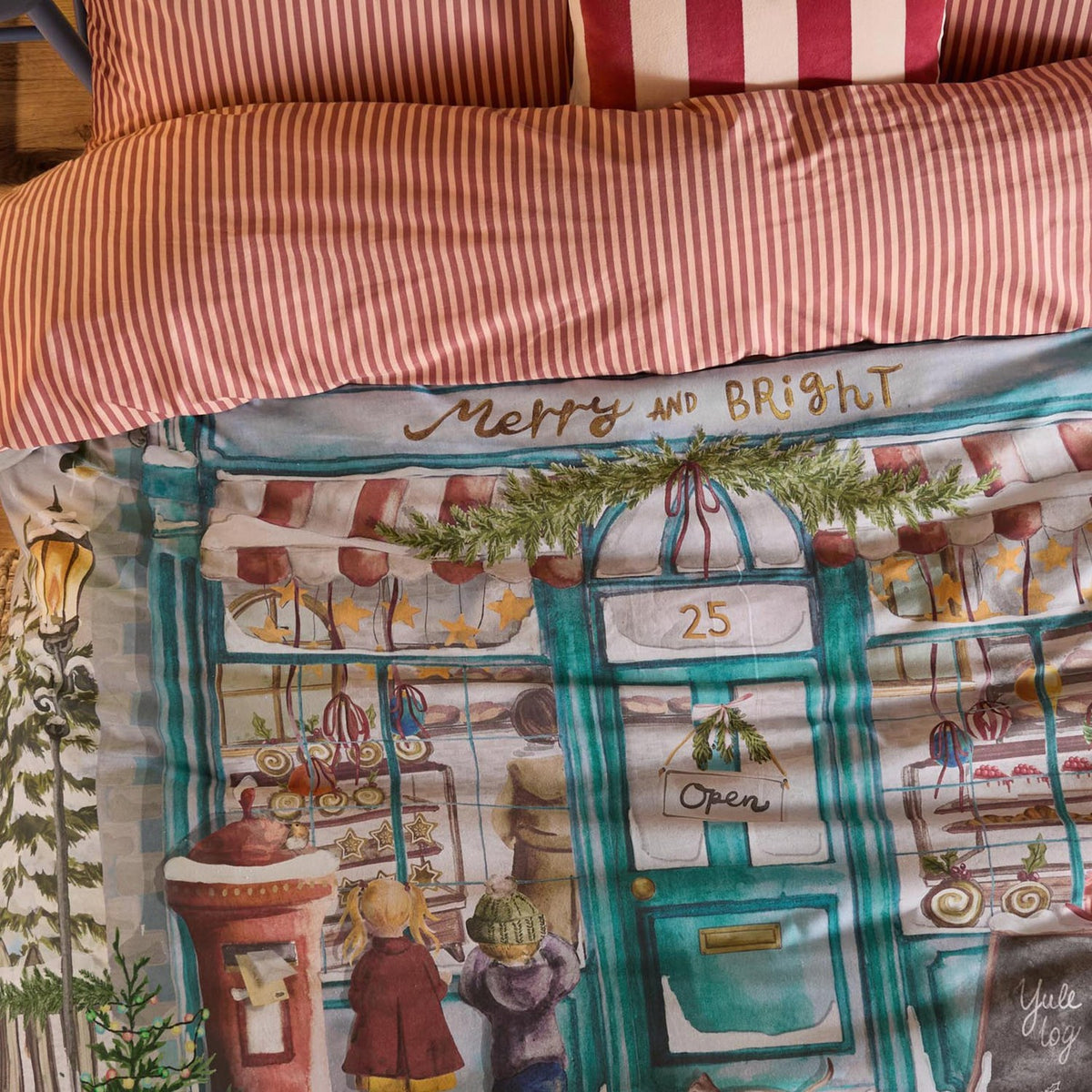 Merry & Bright cotton duvet cover set, showing a red-and-white striped reverse alongside a painted "Merry and Bright" bakery shopfront scene with children, a post box and greenery.