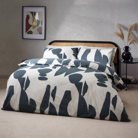 Meta Cotton Abstract Duvet Cover Set in Dusk, featuring slate blue abstract shapes on a cream ground with matching pillowcases, shown on a bed with cane headboard in a bedroom setting.