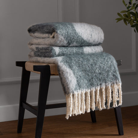 Meta Jacquard Polyester Throw in Dusk, folded on a stool with fringed cream edge, showing a blue-grey abstract pattern on a faux mohair finish.