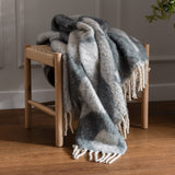 Meta Jacquard Polyester Throw in Dusk, draped over a wooden stool with woven seat, showing grey and cream abstract pattern with fringed edges in a living room setting.
