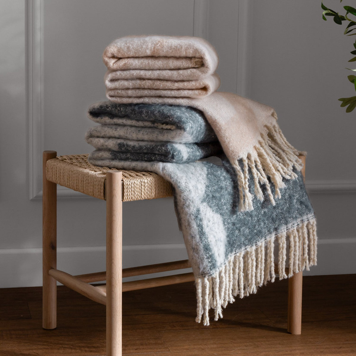 Meta Jacquard Polyester Throw in Dusk, folded with fringed edges in blue-grey abstract pattern on cream, stacked with a beige throw on a woven wooden stool.